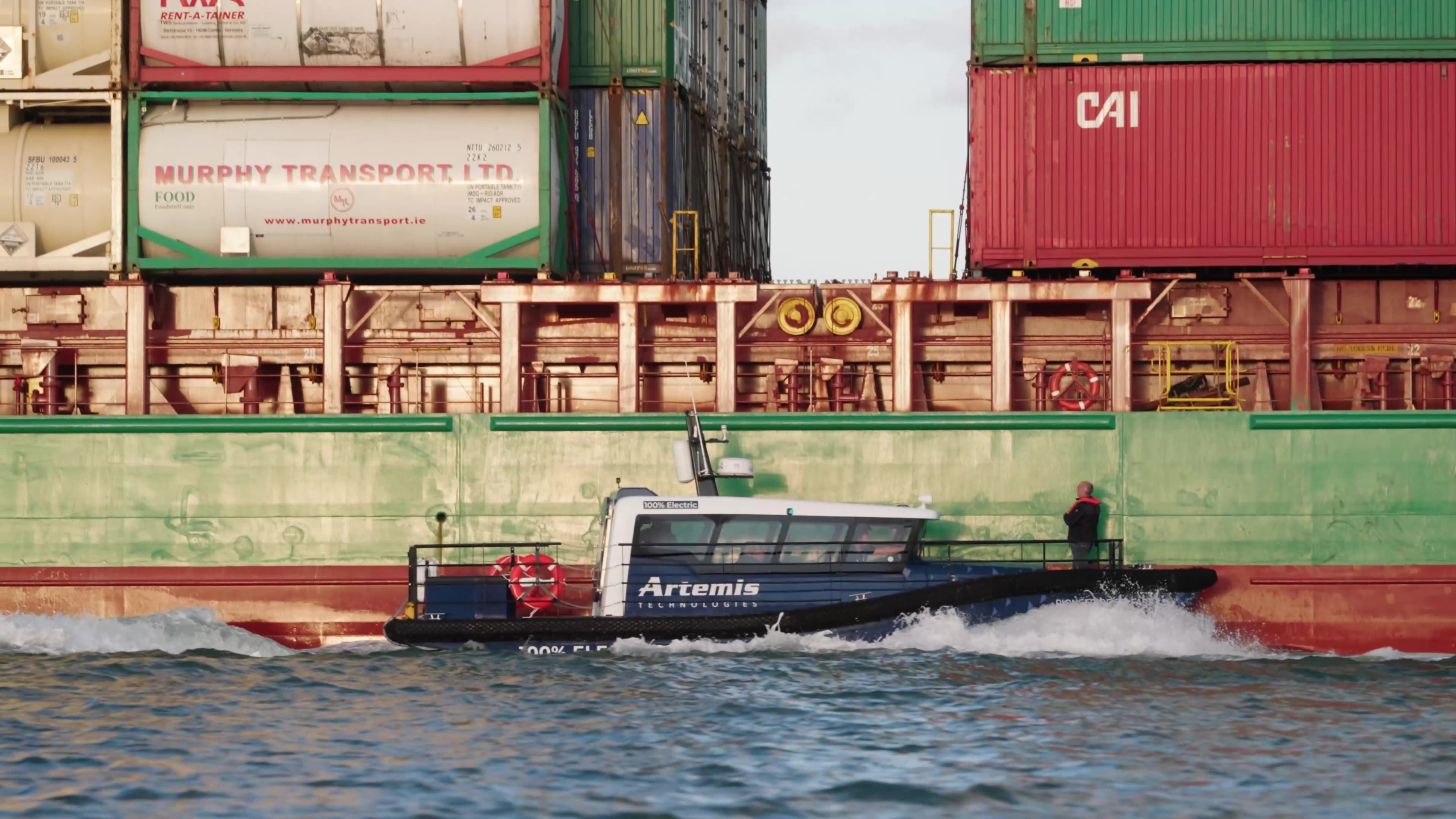 Artemis eFoiler&reg; 12 metre vessel making contact with a large red container ship with a person at the bow of the smaller vessel near to boarding position
