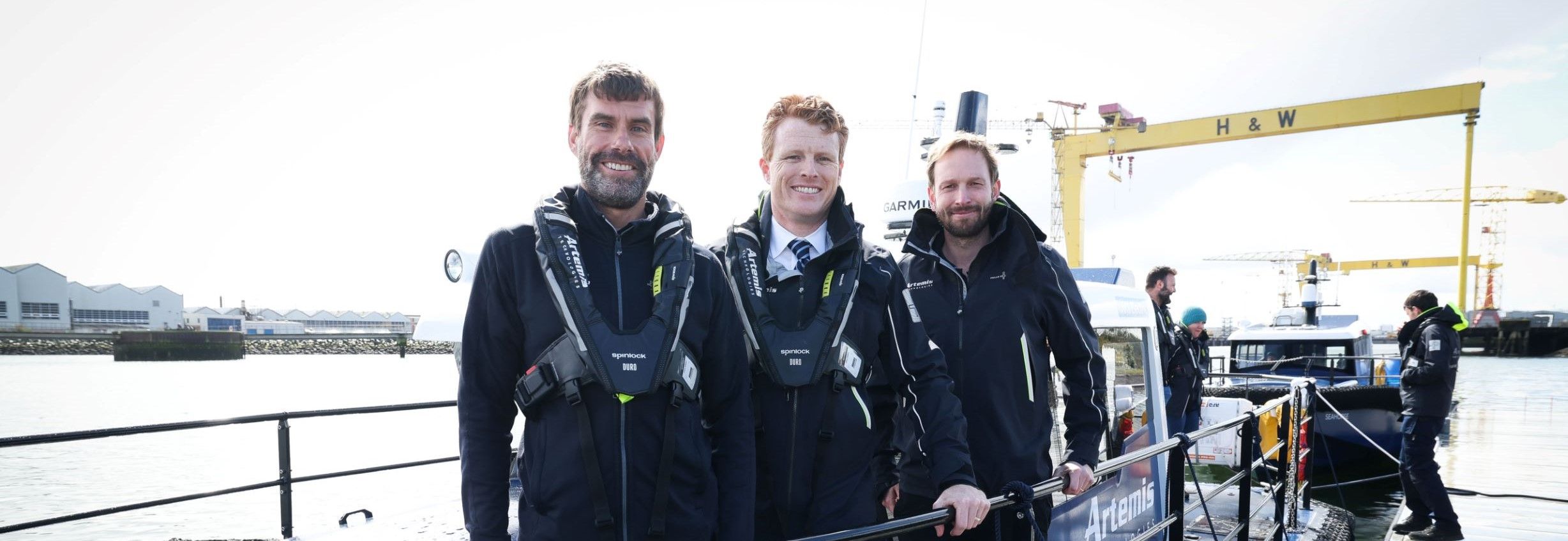 Iain Percy, David Tyler and Joe Kennedy standing on pioneer with harland and wolf cranes in background