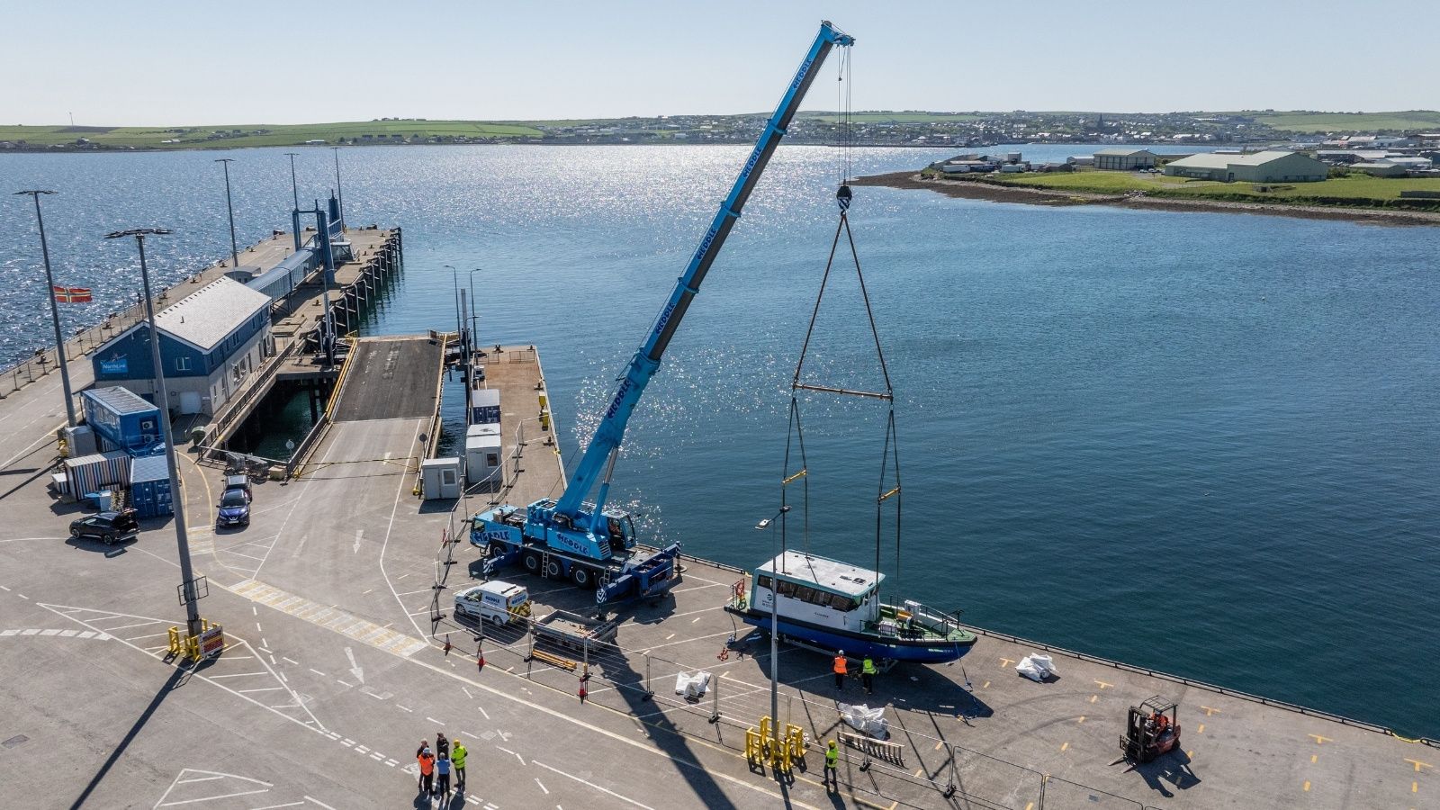 Picture of the Artemis EF-12 Workboat XL being held by a blue crane on a pier in Orkney with the harbour in the background the sun shining on the water and houses in distance.
