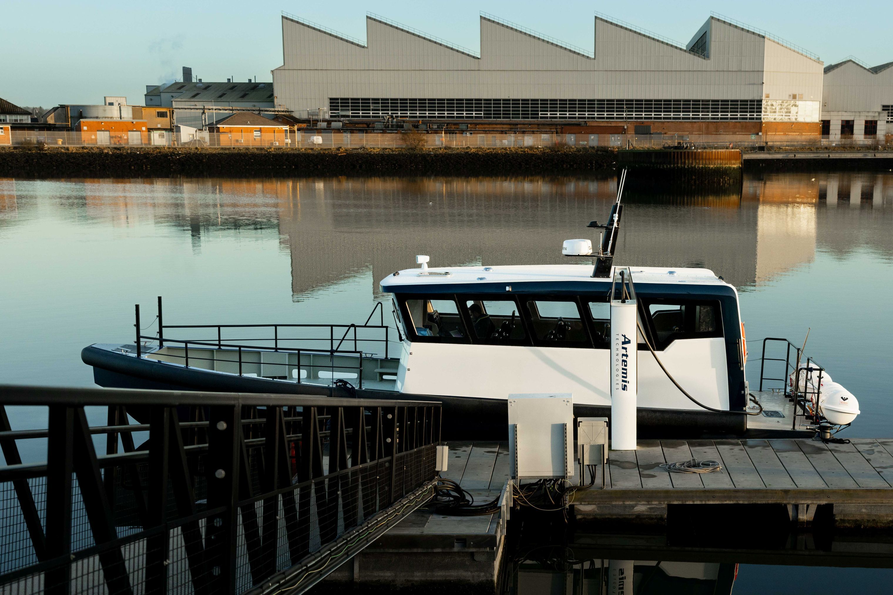 A white and navy 100% electric Artemis EF-12 Workboat XL in the sunlight moored at a pontoon getting charged up by shoreside charger.
