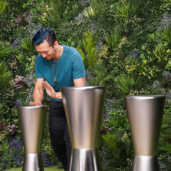 A person with blue hair smiles while playing sleek, metallic outdoor djembe drums in a lush, green garden setting, conveying joy and creativity.