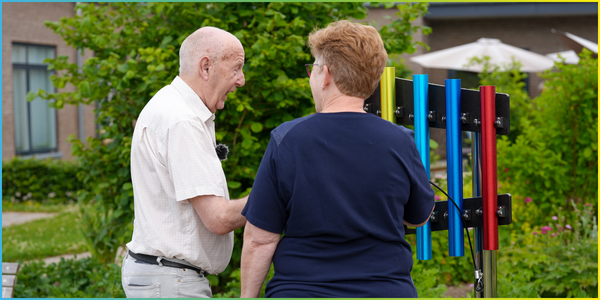 A senior gentlemen with dementia standing and playing a rainbow colored set of outdoor chimes with his carer