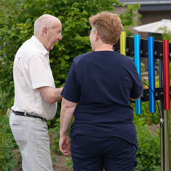 a senior man standing with a healthcare professional playing a set of colourful outdoor musical chimes together in a memory care garden
