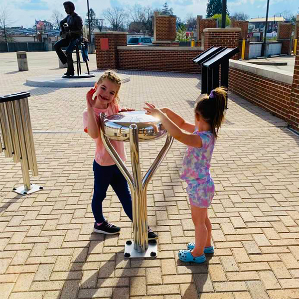 Two young girls having fun on a stainless steel tongue drum at the Bill Withers Memorial Plaza outdoors.