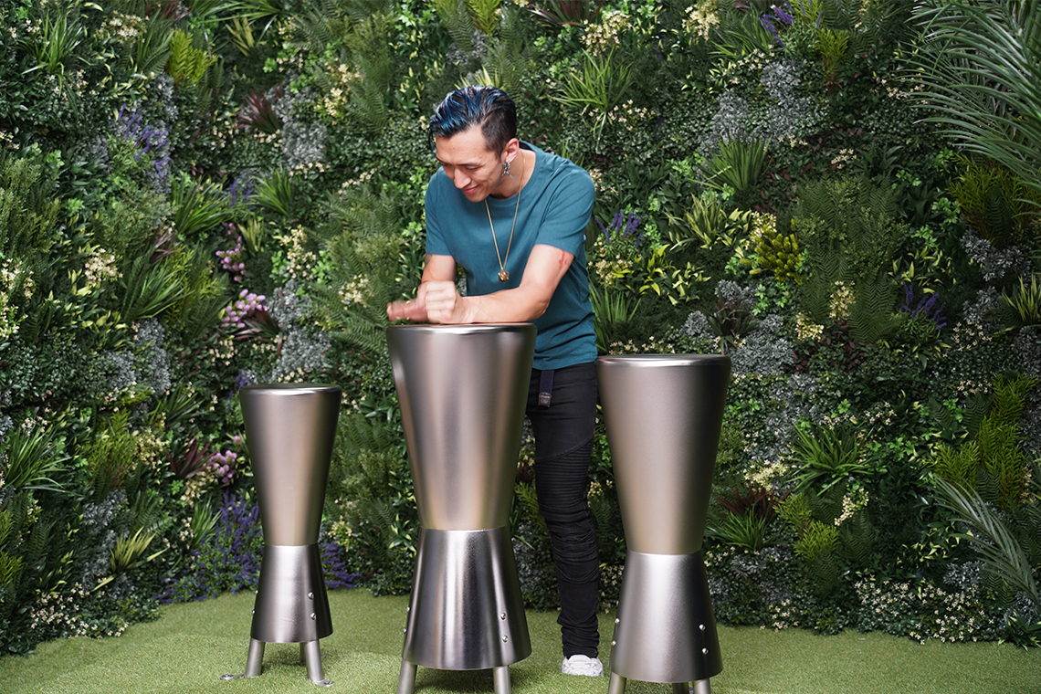 A person with blue hair smiles while playing sleek, metallic outdoor djembe drums in a lush, green garden setting, conveying joy and creativity.