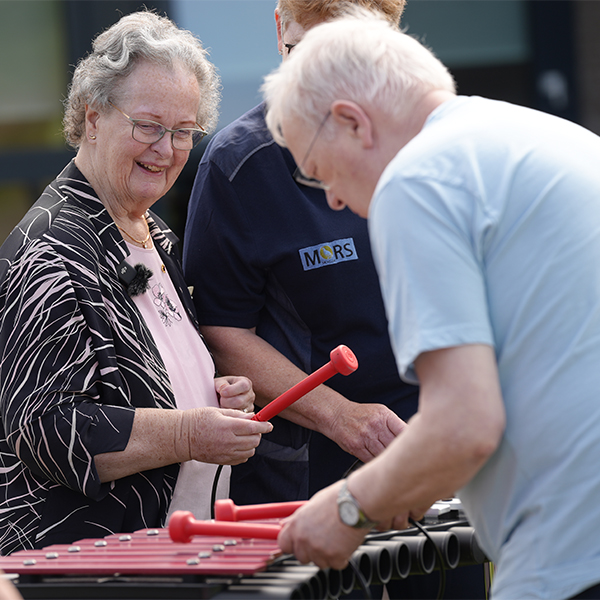 A senior woman and her caregiver happily playing a colorful outdoor xylophone in a serene memory care garden