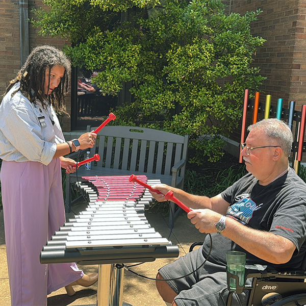 a senior man seated in a wheelchair playing an outdoor musical instrument with a care worker