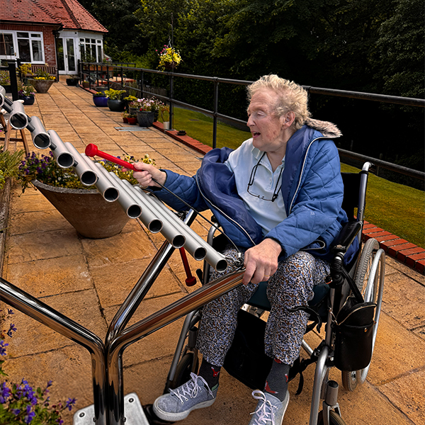 a senior lady in a wheelchair playing a large silver outdoor xylophone in a senior living courtyard setting