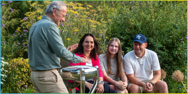 a senior man playing an outdoor drum in a garden with his family sat next to him on a bench watching and smiling