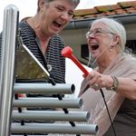 two senior ladys one holding a red mallet playing an outdoor metallophone and laughing