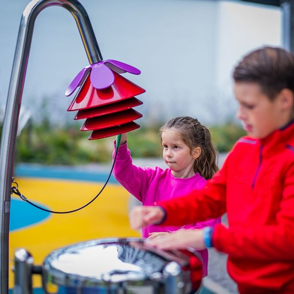 young girl playing a red musical chime shaped as a bell flower with a boy in the foreground out of focus playing a stainless steel tongue drum