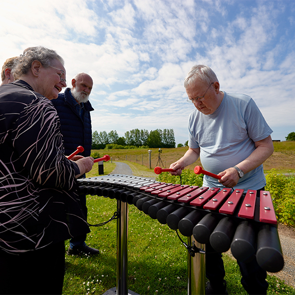 a man in a memory care garden engaged in playing a large outdoor xylophone, surrounded by fellow seniors smiling and watching