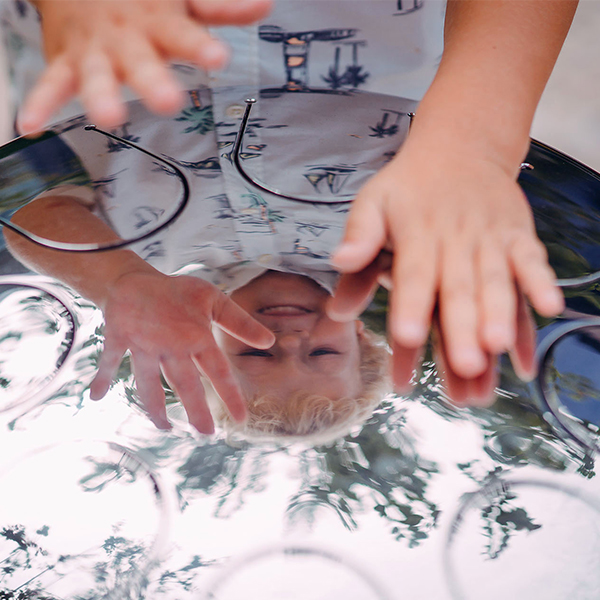 Refelction of a boys face who is playing a stainless steel tongue drum outdoors