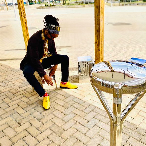 man seated on and playing a metal cajon drum next to a stainless steel outdoor tongue drum at the bill withers memorial plaza