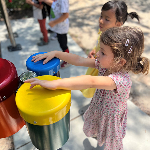 young girl playing a colorful set of three outdoor bongo drum with her hands in a museum garden