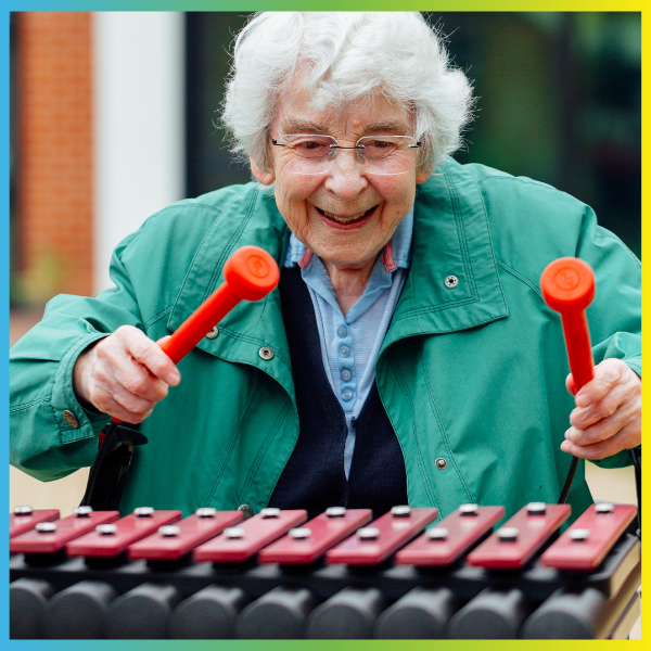 a senior lady holding two red mallets and playing an outdoor xylophone enthusiastically 