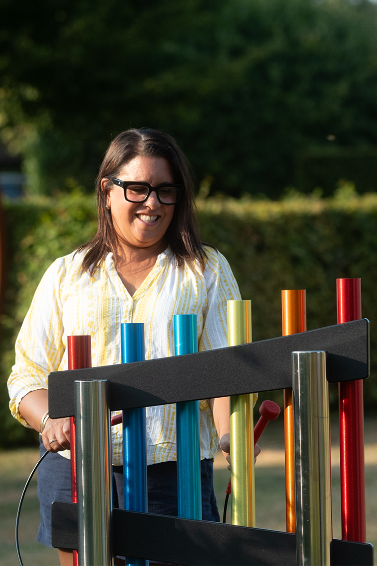 an adult lady standing in from of a set of rainbow chimes in a summer garden