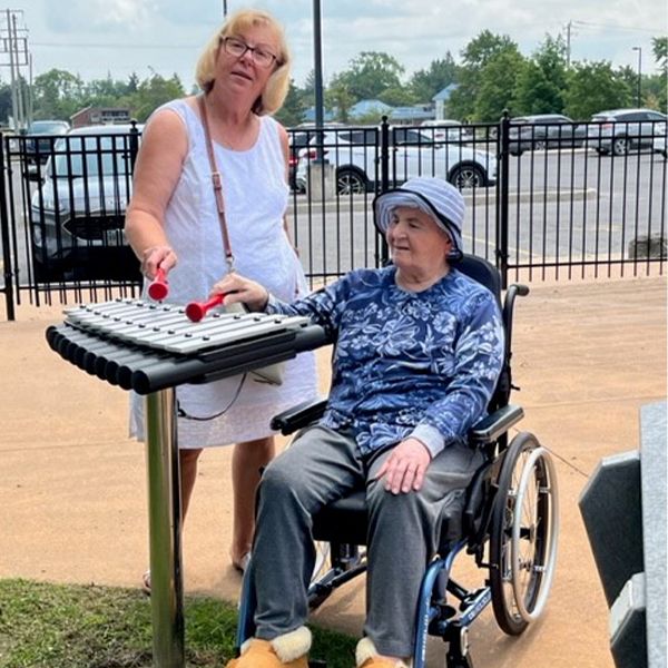 two ladies one standing and one in a wheelchair playing an outdoor musical instrument called a cadenza