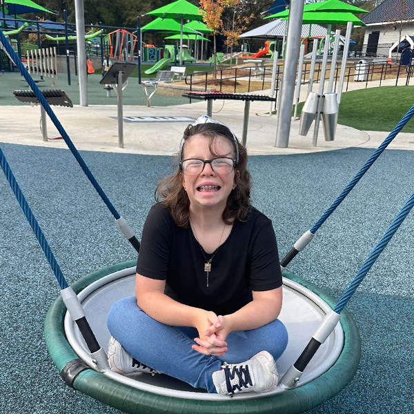 a teenage girl in a swing hammock in front of outdoor musical instruments in an inclusive play park