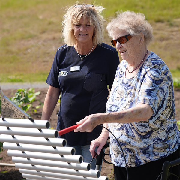 A senior woman stands in a garden, playing an outdoor musical instrument for her carer, creating a joyful moment together.