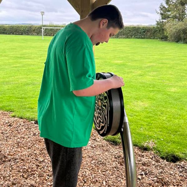A young man with autism happily playing on a large circular black and silver rain wheel at the Thomas Centre's new music area, surrounded by various other outdoor musical instruments.