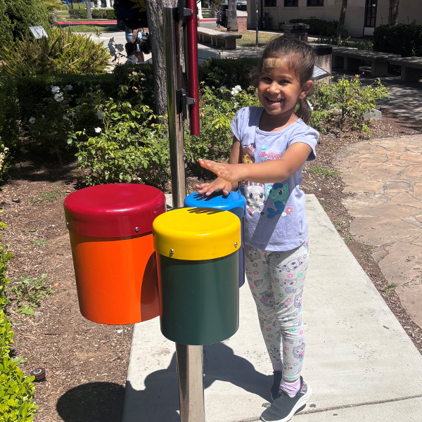 a young girl smiling and playing a set of outdoor bongo drums with her hands in a childrens museum garden