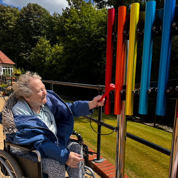 a senior lady seated in a wheelchair playing a set of colorful outdoor musical chimes in a senior living courtyard