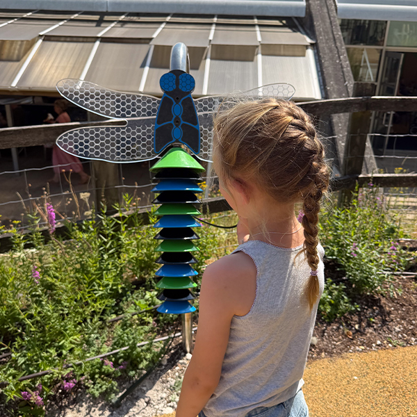 the back of a young girl playing an outdoor musical instrument shaped like a dragonfly with bells in green, blue and black as its tail