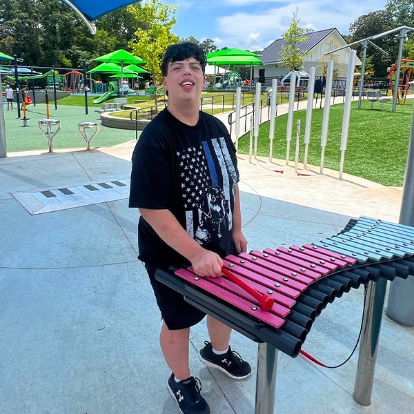 a young man under a shade sail playing an outdoor xylophone in an inclusive play park