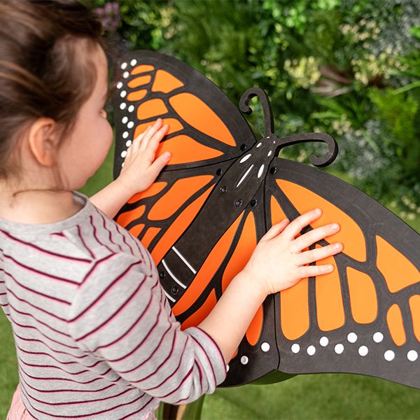 A child interacts with a large butterfly music sculpture, featuring vivid orange and black wings, in a lush garden setting.