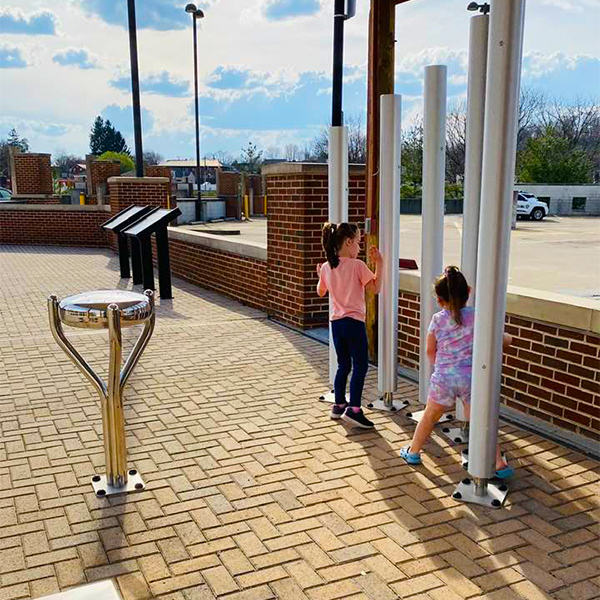 Two children playing on a brick walkway, interacting with tall metal outdoor chimes at the Bill Withers Memorial Plaza.