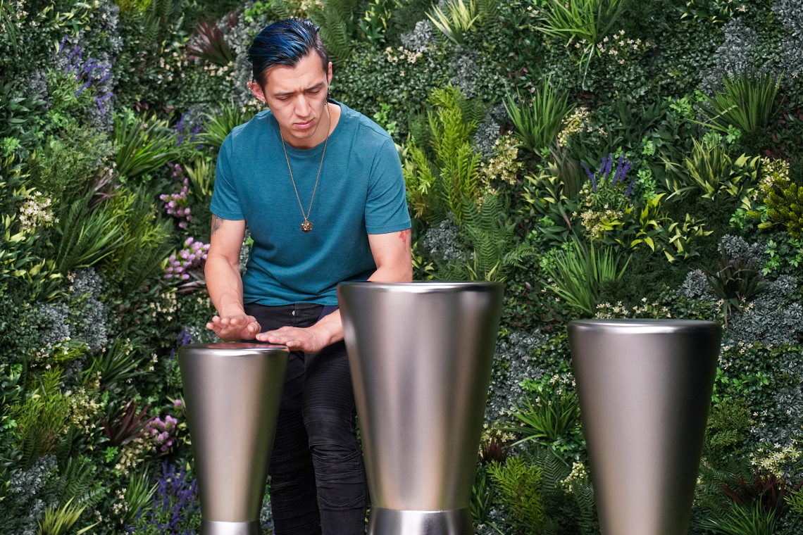 A person with blue hair smiles while playing sleek, metallic outdoor drumsin a lush, green garden setting, conveying joy and creativity.