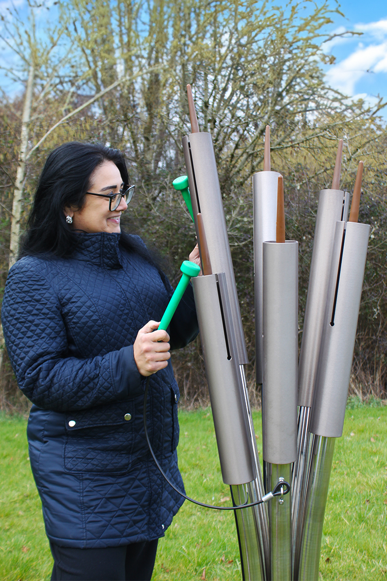 A person in a quilted coat plays cattail chimes, an outdoor musical instrument with aluminum pipes and stainless steel posts. They hold a green pair of mallets against the backdrop of trees and a cloudy sky.