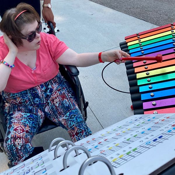 a teenage girl in a wheelchair playing a rainbow colored outdoor xylophone in a music park at summer camp